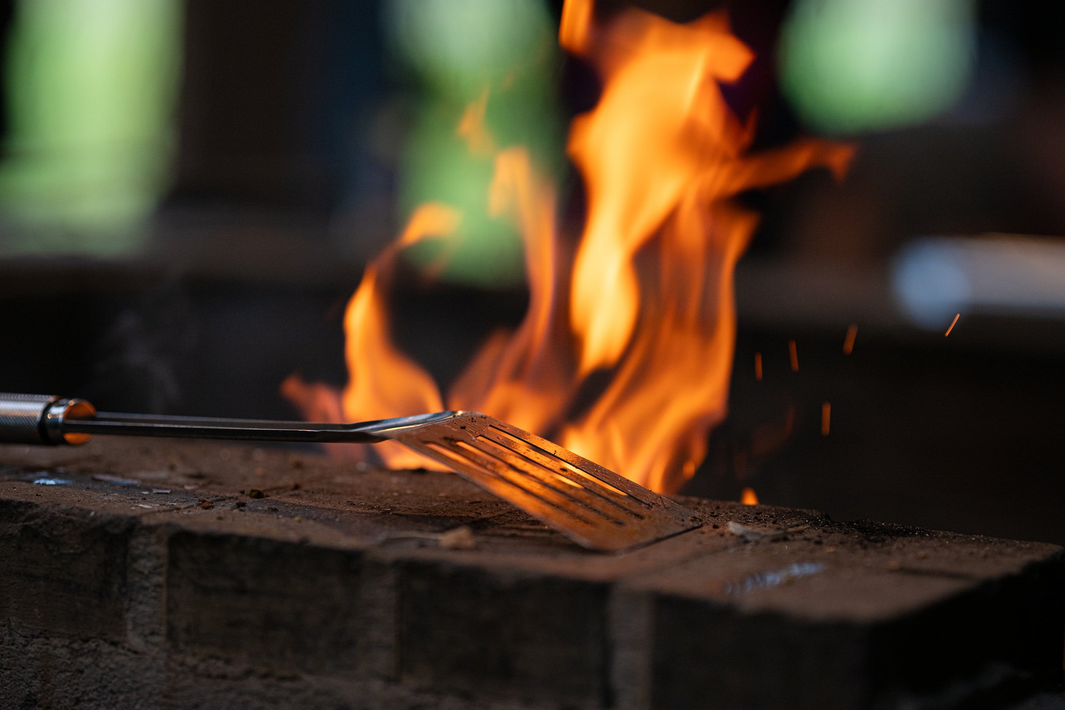 Vorbereitung für den GrillGorilla Einsatz: Nahaufnahme eines Grills mit loderndem Feuer und Grillwender auf einer Steinmauer.